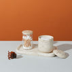 A still life of a beige stone tray holding a lidded jar with cotton pads, a lidded jar with cotton swabs, and a brown hair claw clip on a light blue surface, with an orange backdrop.