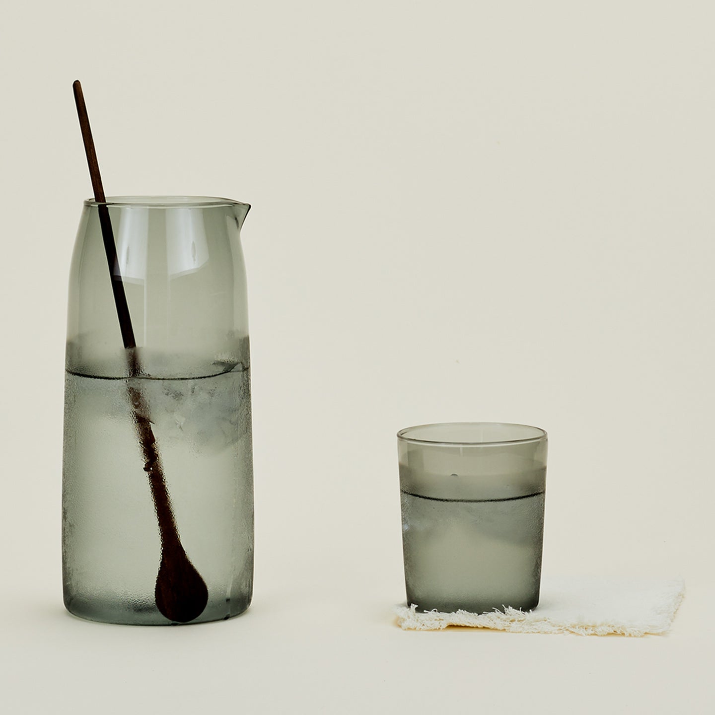 Essential Glassware Pitcher in Smoke, filled with ice water, along with a drinking glass and stirring spoon.